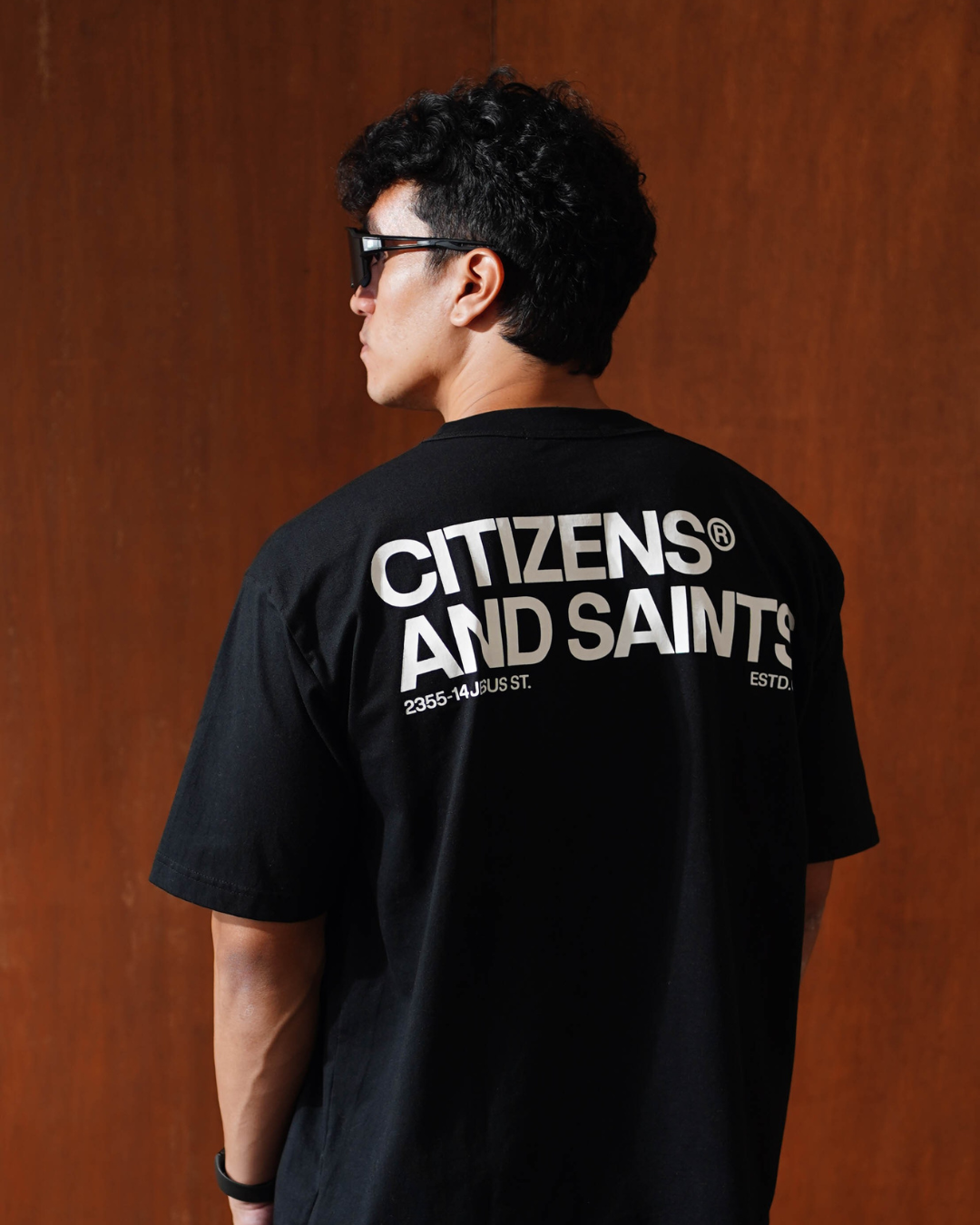 Person wearing a black 'Citizens and Saints' t-shirt against a brown background